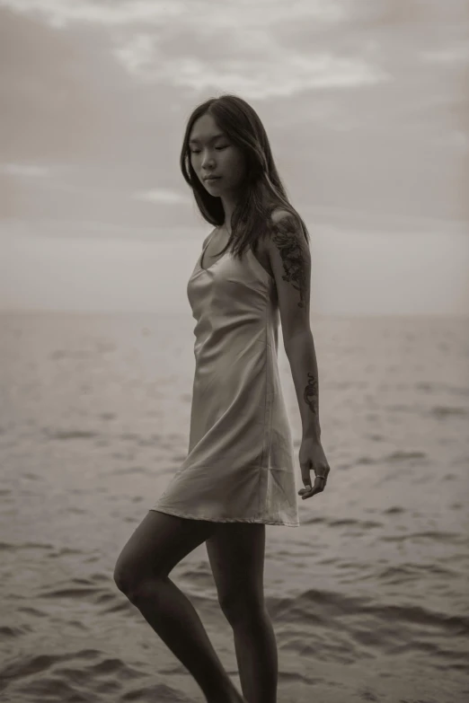 a woman standing on top of a beach next to the ocean, a black and white photo, unsplash, renaissance, wearing a wet white short dress, of taiwanese girl with tattoos, wearing a nightgown, wearing a camisole