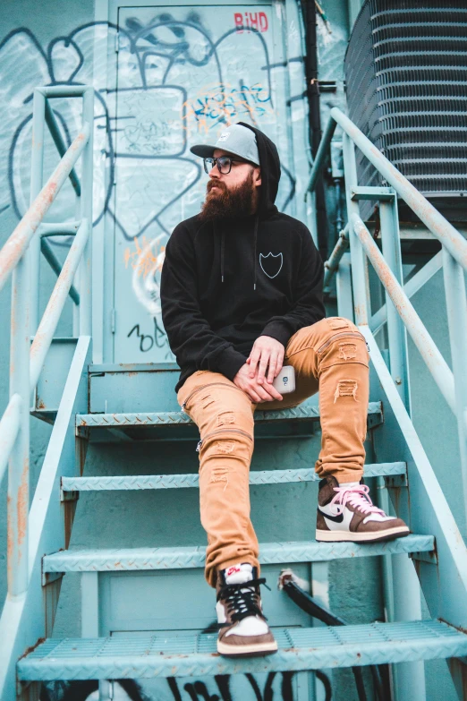 a man sitting on top of a set of stairs, inspired by Drew Tucker, pexels contest winner, lyco art, thick brown beard, trending on r/streetwear, wearing jeans and a black hoodie, vivid and detailed