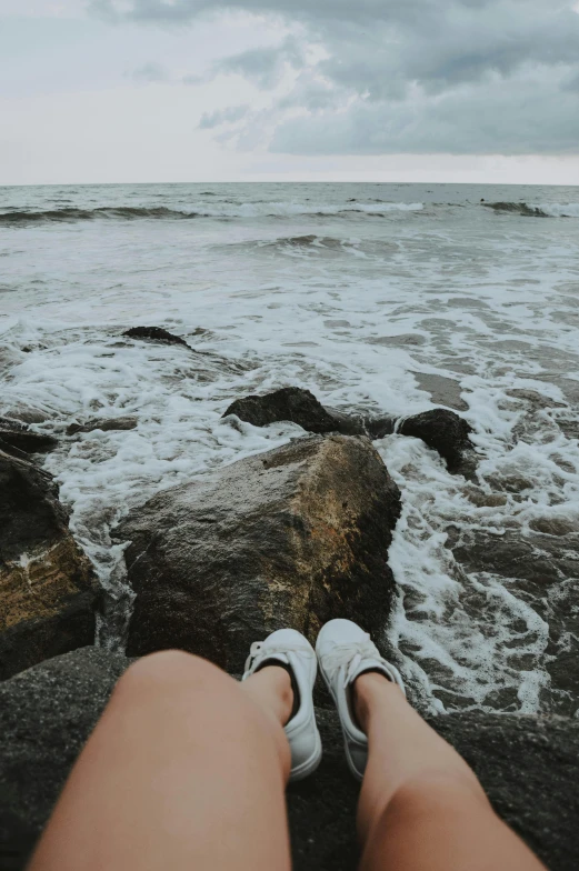 a person sitting on a rock next to the ocean, wet feet in water, wearing white sneakers, turbulence, festivals