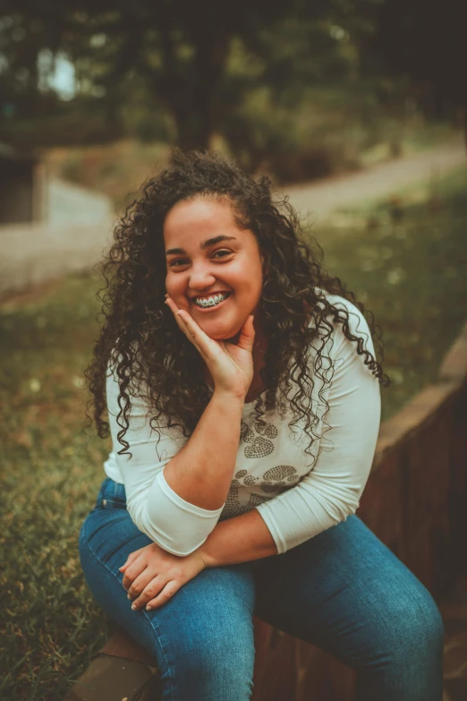 a woman with curly hair sitting on a wall, by Almada Negreiros, pexels contest winner, happily smiling at the camera, full figured, in a park, slightly pixelated