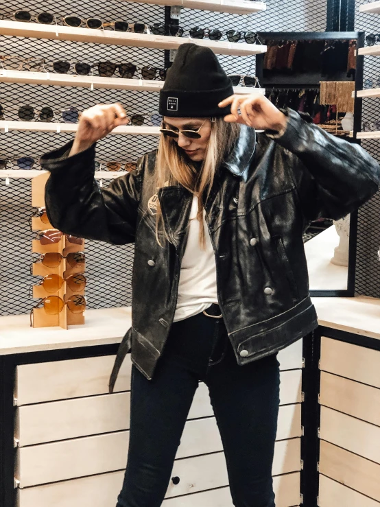 a woman standing in front of a counter in a store, an album cover, by Robbie Trevino, trending on unsplash, she wears leather jacket, wearing sunglasses and a cap, satisfied pose, view from the side