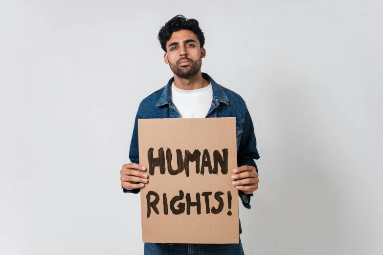 a man holding a sign that says human rights, an album cover, trending on pexels, renaissance, indian, background image, cardboard cutout, young adult male