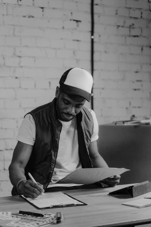 a man sitting at a table in front of a laptop, a black and white photo, inspired by Theo Constanté, pexels contest winner, process art, handsome hip hop young black man, writing on a clipboard, artist wearing overalls, promotional image