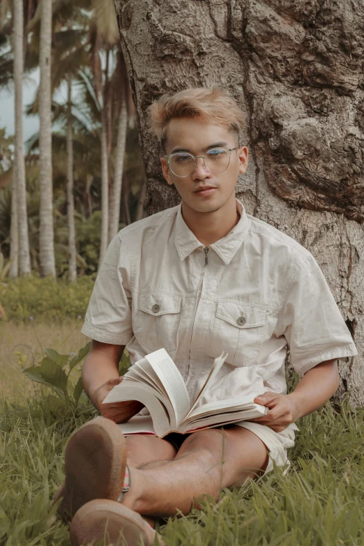 a man sitting under a tree reading a book, by Robbie Trevino, androgynous face, transparent glasses, 2019 trending photo, twink