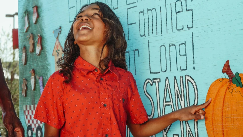 a woman standing in front of a wall with a painted pumpkin on it, an album cover, pexels contest winner, red shirt, happy kid, aboriginal australian hipster, looking upwards