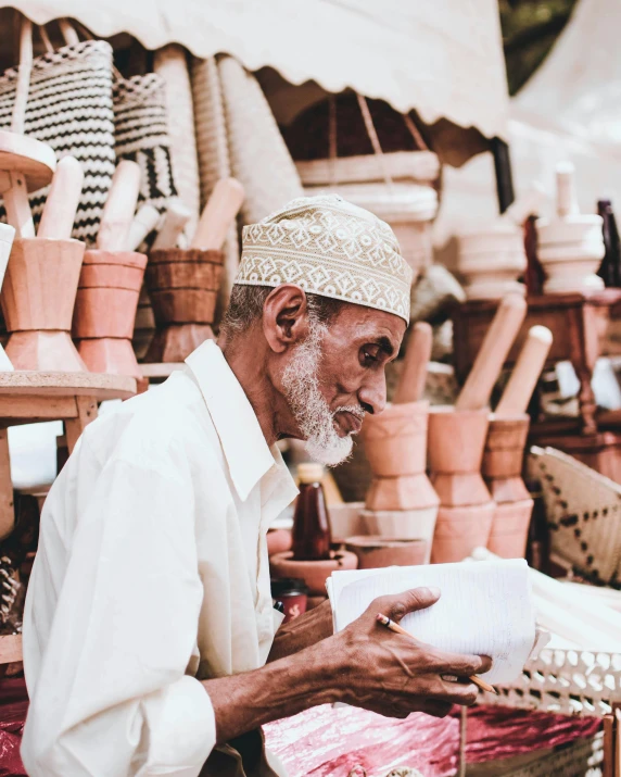 a man sitting in front of a display of pottery, by Ismail Acar, pexels contest winner, reading the book about love, wearing a white bathing cap, somali attire, woodturning