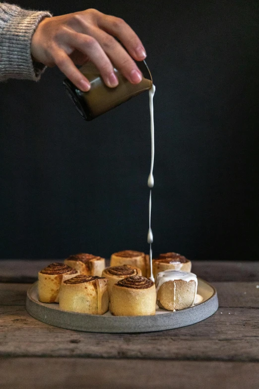 a person pouring icing onto a plate of cinnamon rolls, by Susy Pilgrim Waters, sydney, medium format, raku, grey