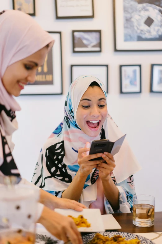 a group of women sitting around a table looking at a cell phone, a picture, by Maryam Hashemi, pexels contest winner, hurufiyya, mutahar laughing, wearing a head scarf, photoshoot for skincare brand, medium close-up shot