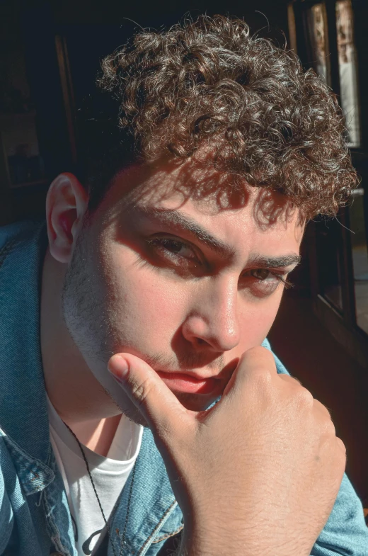 a man sitting at a table with his hand on his chin, an album cover, by Leo Michelson, trending on pexels, curly bangs, blue symmetric eyes 24yo, headshot profile picture, in the sun