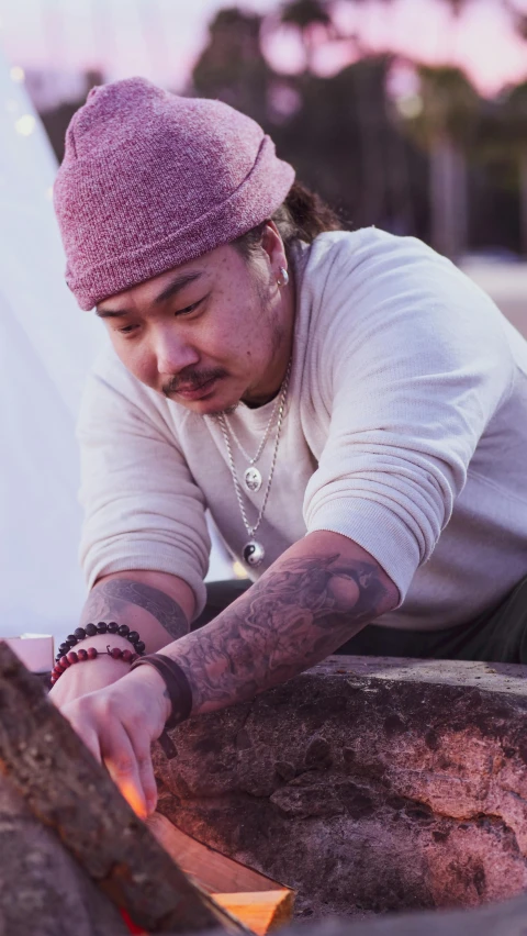 a man sitting on top of a rock next to a fire, a tattoo, inspired by I Ketut Soki, trending on unsplash, process art, small chin, vintage color, professional woodcarving, chris chan