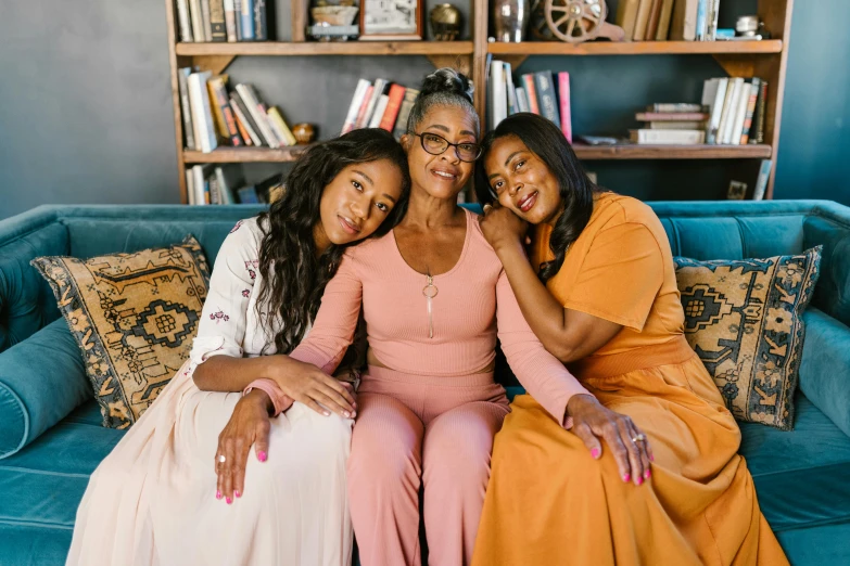 three women sitting on a couch in front of a bookshelf, a portrait, by Carey Morris, pexels, african american woman, brown and pink color scheme, motherly, embracing