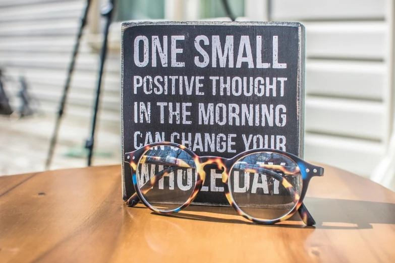 a pair of glasses sitting on top of a wooden table, by Arabella Rankin, unsplash, private press, motivational poster, small smile, sign, full daylight
