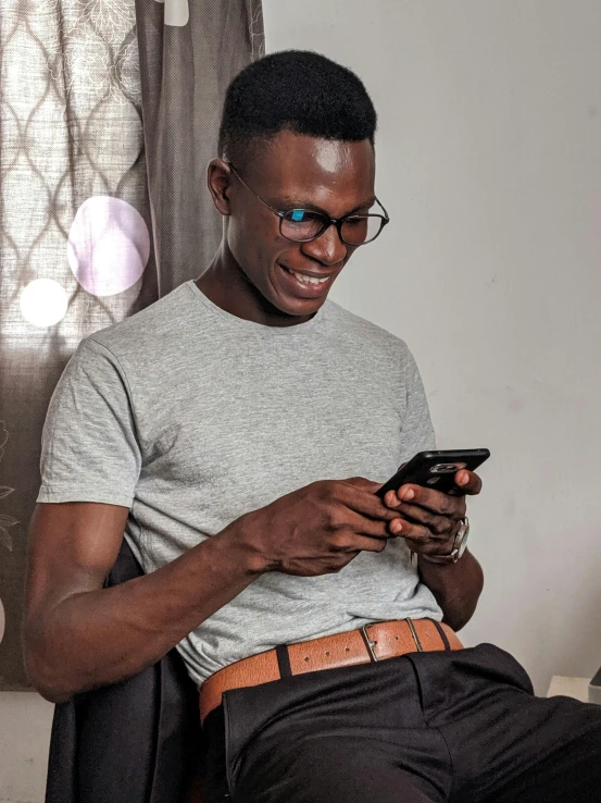 a man sitting in a chair looking at his cell phone, by Chinwe Chukwuogo-Roy, trending on pexels, wearing reading glasses, with grey skin, wearing tight shirt, he is about 20 years old | short