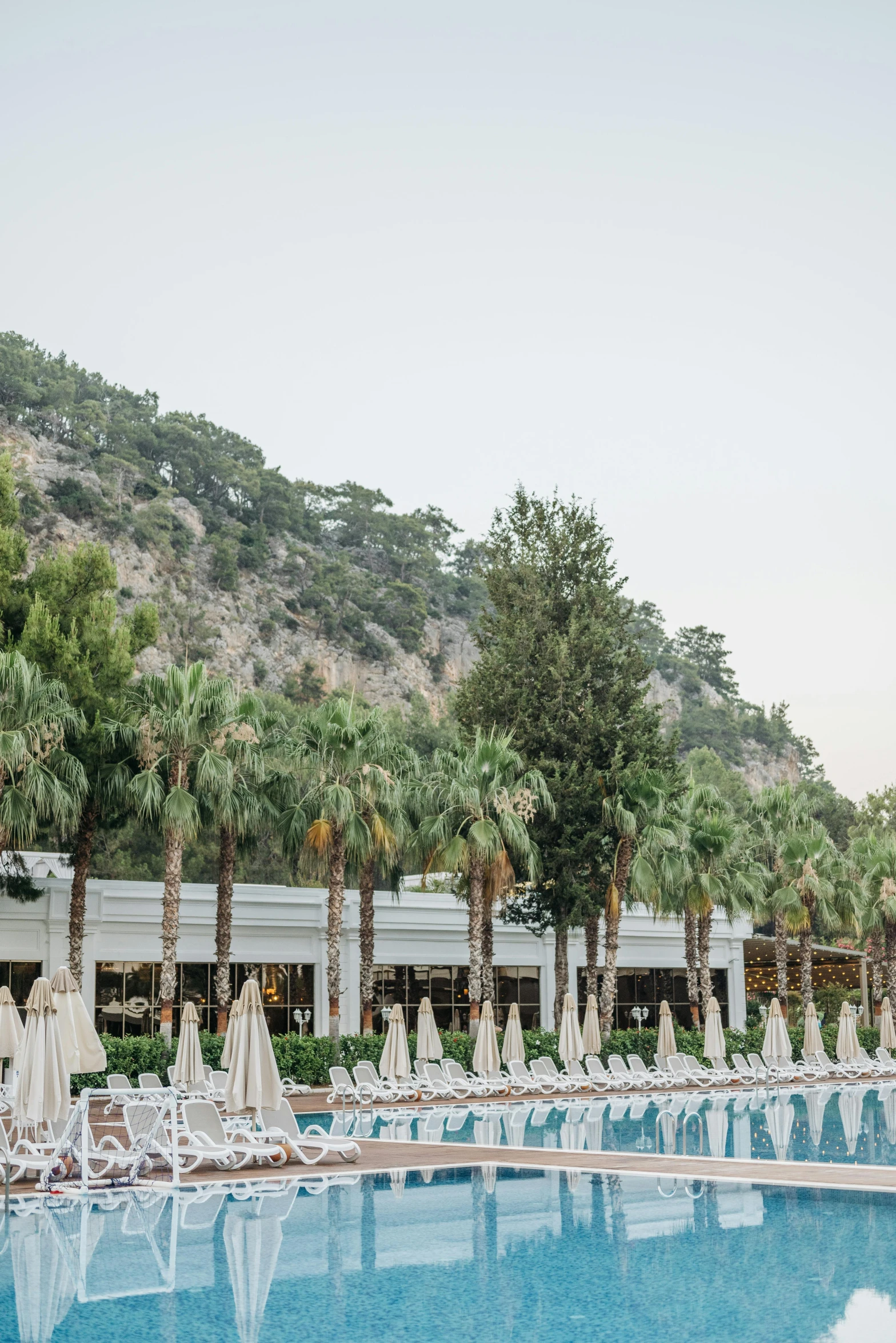 an outdoor swimming pool with lounge chairs and umbrellas, hurufiyya, trees and cliffs, jen atkin, kids, turkish and russian