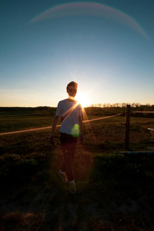 a person standing in front of a fence at sunset, 5k, in a field, sun overhead, dylan kowalski