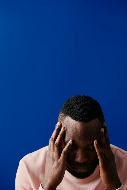 a man sitting at a table with his head in his hands, an album cover, pexels, blue backdrop, ( ( dark skin ) ), stressed, ignant