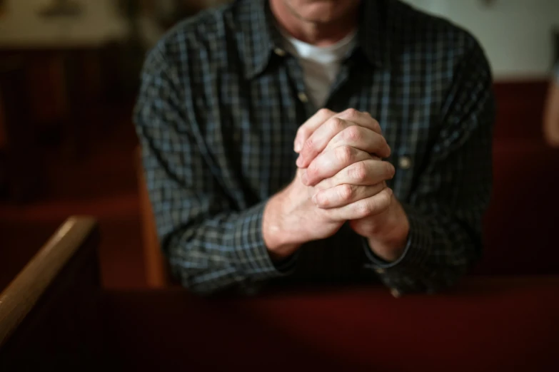 a man sitting in a church holding his hands together, a portrait, by Dan Frazier, pexels, hand on table, middle aged man, plain background, multiple stories