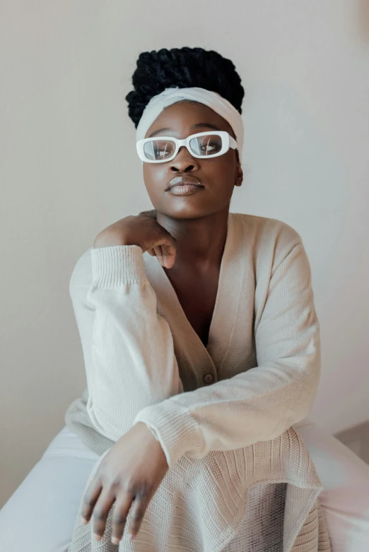 a woman with glasses sitting on a bed, by Dulah Marie Evans, trending on unsplash, afrofuturism, white outfit, square rimmed glasses, lupita nyong'o, shaven