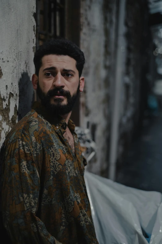 a man with a beard leaning against a wall, an album cover, by Alejandro Obregón, pexels contest winner, real life photo of a syrian man, in a movie still cinematic, lgbtq, ( ( theatrical ) )