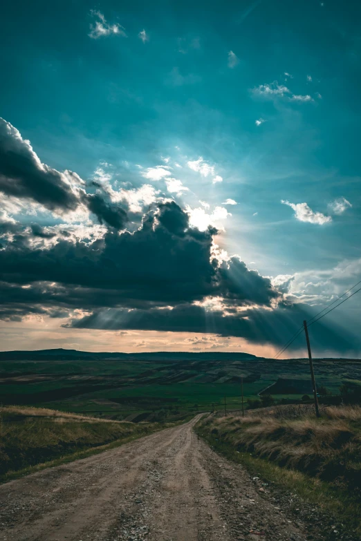 a dirt road in the middle of a field under a cloudy sky, a picture, pexels contest winner, renaissance, sun streaming in, teal sky, overlooking, dramatic lightin