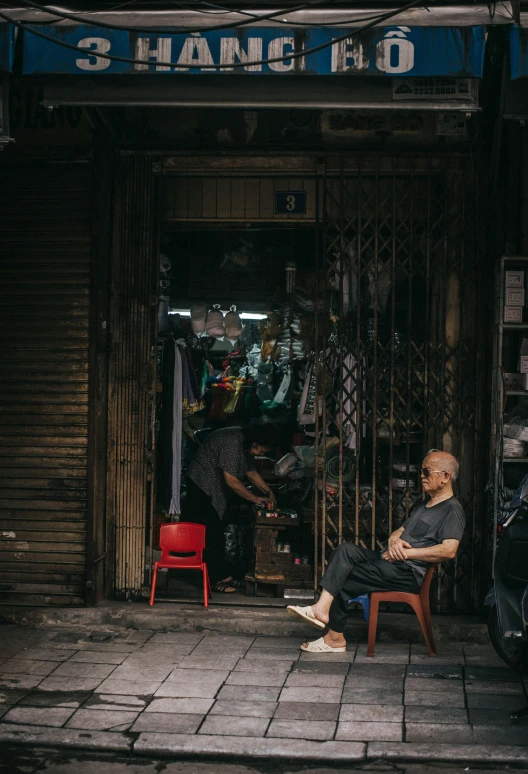 a man sitting on a chair in front of a store, pexels contest winner, jia ruan, bald man, ben lo, markets