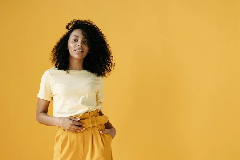 a woman standing in front of a yellow wall, wearing pants and a t-shirt, brown skinned, wavy hair yellow theme, portrait featured on unsplash