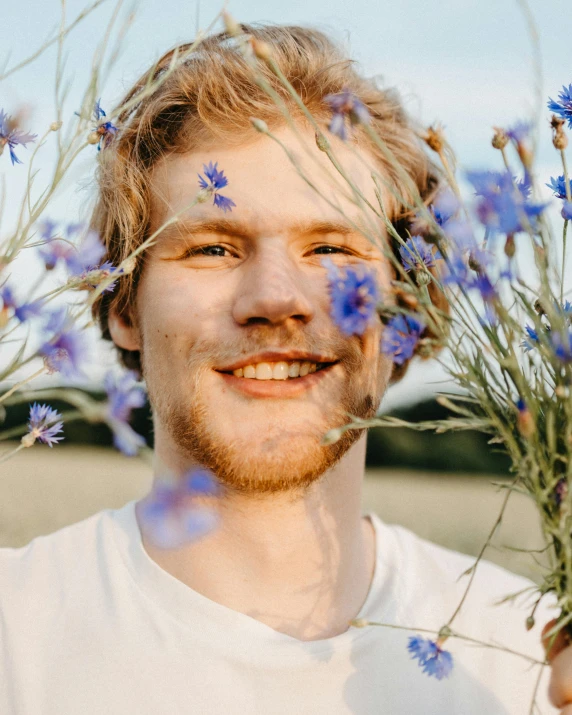 a man holding a bunch of flowers in front of his face, a colorized photo, trending on unsplash, naturalism, ginger hair with freckles, grinning lasciviously, blue flowers bloomed all over, charming sly smile