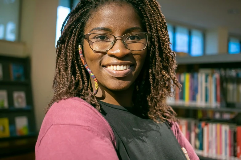 a woman with glasses standing in front of a bookshelf, by Stokely Webster, smiling young woman, atiba jefferson, close up portrait photo, thumbnail