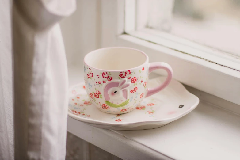 a cup and saucer sitting on a window sill, inspired by Annabel Kidston, pink, rabbit, small, multi - coloured