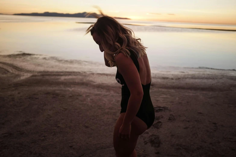 a woman standing on a beach next to a body of water, a picture, profile image, warm glow, is wearing a swimsuit, skye meaker