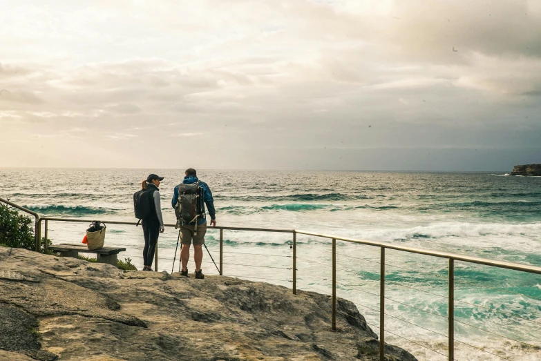 a couple of people standing on top of a cliff next to the ocean, by Peter Churcher, people angling at the edge, manly, 4k photo”, conde nast traveler photo