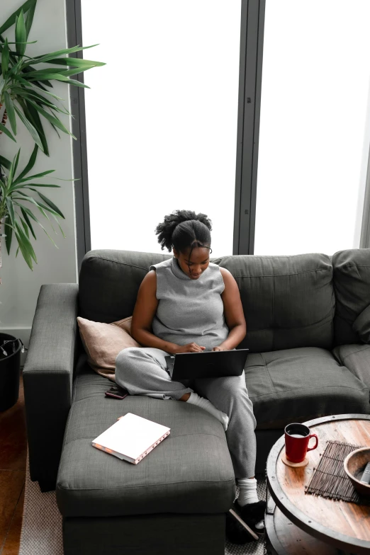 a woman sitting on a couch using a laptop, by Carey Morris, pexels contest winner, ( ( dark skin ) ), high angle, full body profile, serious business