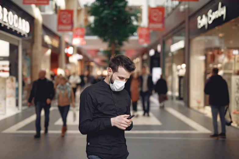 a man wearing a face mask while looking at his phone, trending on pexels, mall background, people walking around, avatar image, dust mask