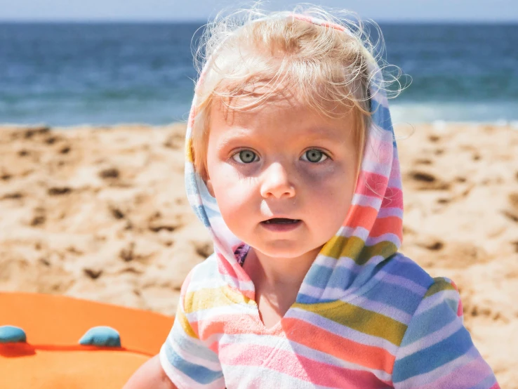 a little girl standing on top of a sandy beach, wearing a towel, full of colour, staring into the camera, liam brazier