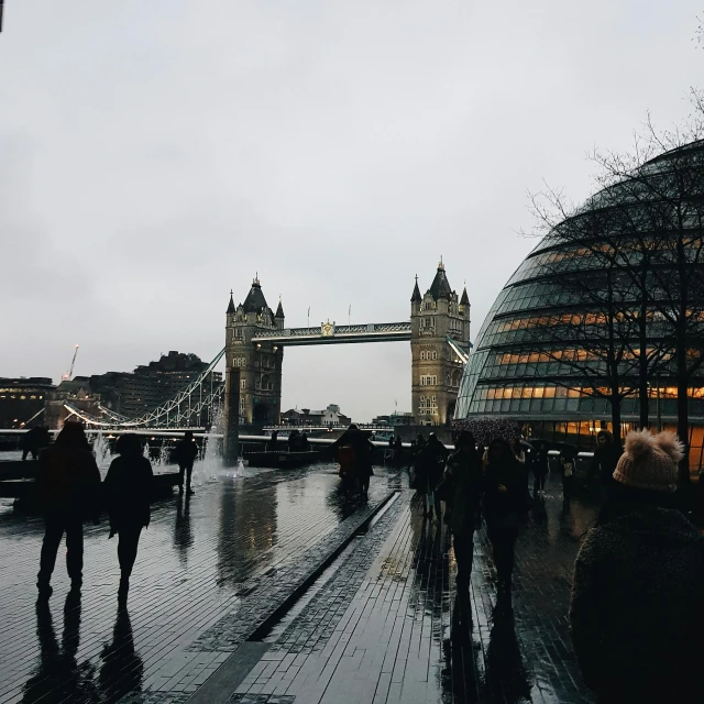 a group of people walking across a wet street, by Emma Andijewska, pexels contest winner, modernism, tower bridge, gray sky, massive arch, view from the side”