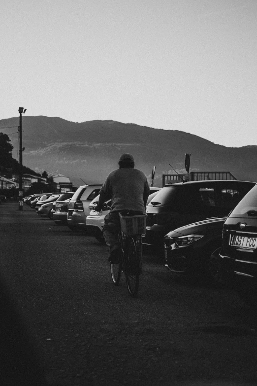 a man riding a bike down a street next to parked cars, a black and white photo, mountain, late summer evening, seaside, lowres