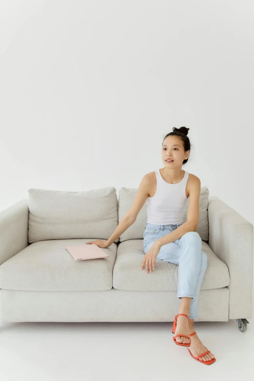 a woman sitting on a couch with a laptop, inspired by Fei Danxu, pexels contest winner, minimalism, pastel clothing, excited, wearing jeans, off - white collection