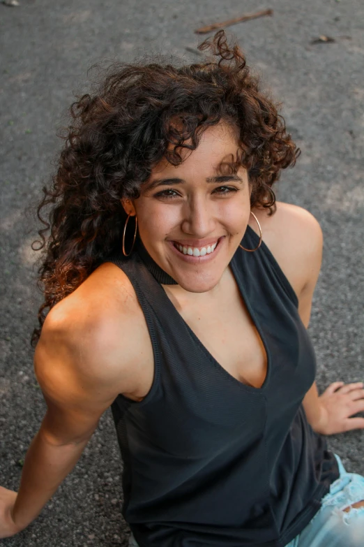 a woman sitting on the ground next to a fire hydrant, an album cover, by Jessie Algie, pexels contest winner, happening, dark short curly hair smiling, she is wearing a black tank top, high angle closeup portrait, ana de la reguera portrait