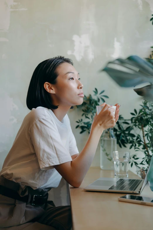 a woman sitting at a table in front of a laptop, inspired by Ruth Jên, trending on unsplash, realism, set on singaporean aesthetic, pondering, wearing a light shirt, admiring her own reflection