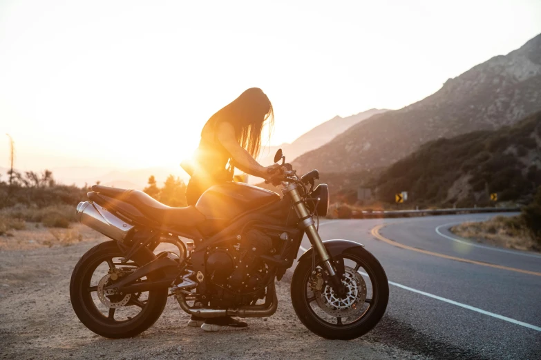 a person sitting on a motorcycle on the side of a road, by Lee Loughridge, trending on unsplash, golden sunlight, woman, triumph, 🚿🗝📝