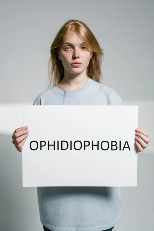 a woman holding a sign that says ophidphobia, shutterstock, antipodeans, on a gray background, atrophy, sadie sink, ohio