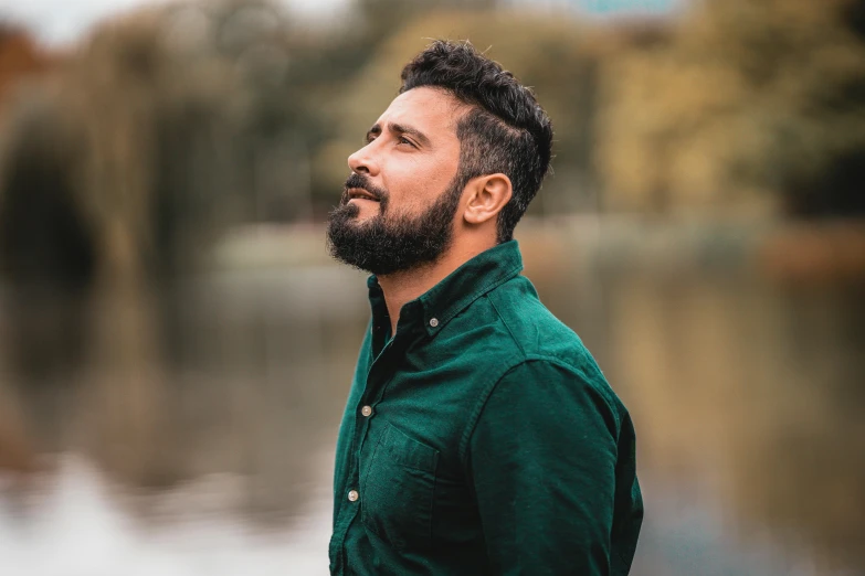 a man standing in front of a body of water, an album cover, pexels contest winner, hurufiyya, very attractive man with beard, profile image, wearing green, confident relaxed pose