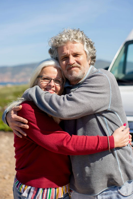 a man and woman hugging in front of a van, gray haired, on the coast, warm friendly expression, gray beard