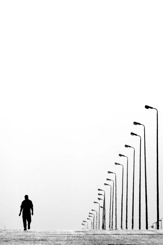 a black and white photo of a person walking down a street, by Zsolt Bodoni, unsplash contest winner, postminimalism, lamps, ffffound, white sky, single silhouette figure