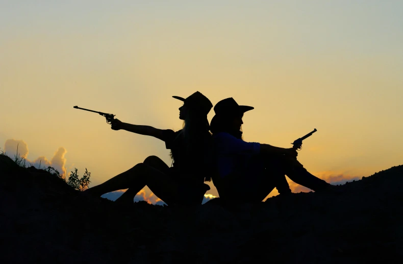 a couple of people sitting on top of a hill, by Linda Sutton, pexels contest winner, holding a shotgun, female cowgirl, evening time, best friends