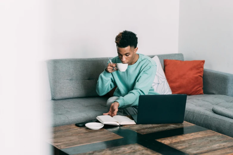 a man sitting on a couch using a laptop, trending on pexels, with teal clothes, table in front with a cup, young commoner, ashteroth