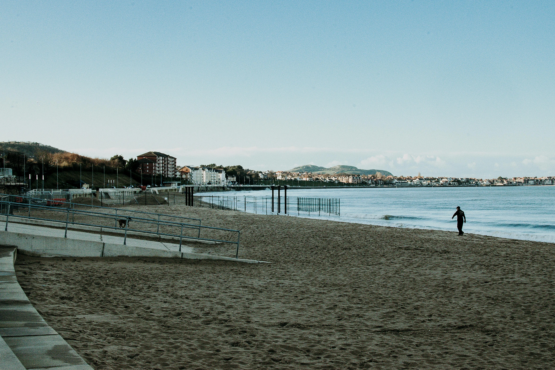 a person standing on a beach next to a body of water, people walking around, building in the distance, spanish, profile image