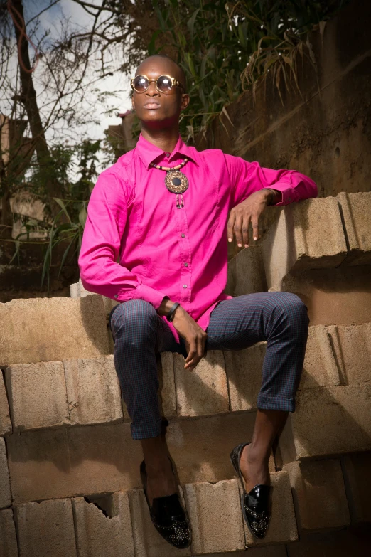 a man in a pink shirt sitting on a wall, by Chinwe Chukwuogo-Roy, pexels contest winner, renaissance, magenta shirt, wears a egyptian ankh necklace, ( ( dark skin ) ), male teenager