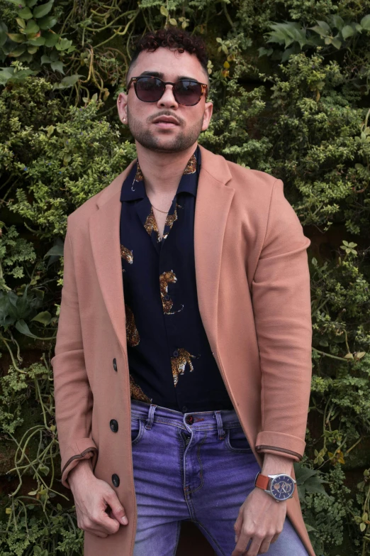 a man standing in front of a bush with his hands in his pockets, an album cover, by Robbie Trevino, trending on pexels, wearing a purple detailed coat, wearing orange sunglasses, singer maluma, headshot profile picture