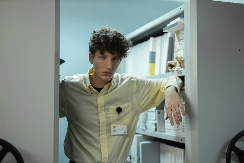 a man in a yellow shirt leaning against a wall, an album cover, by Jacob Toorenvliet, standing in a server room, curly haired, wearing lab coat and a blouse, concerned